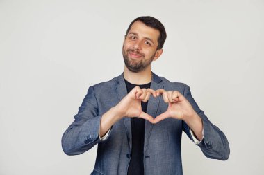 Ceketli, sakallı, gülümseyen genç bir işadamı. Parmaklarıyla kalp şeklinde bir sembol yapıyor. Aşkı ve olumlu romantik duyguyu ifade ediyor. Gri arka planda bir adamın portresi.