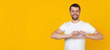 Yakışıklı genç bir adam, beyaz tişörtlü sakallı bir adam, gülümsüyor ve parmağıyla kalp şeklinde bir sembol yapıyor, aşkı ve olumlu bir romantik duyguyu ifade ediyor. Sarı arkaplan