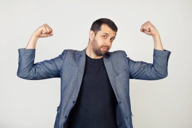 Ceketli, sakallı, kol kaslarını gösteren, gururla gülümseyen genç bir işadamı. Fitness konsepti. Gri arka planda bir adamın portresi.