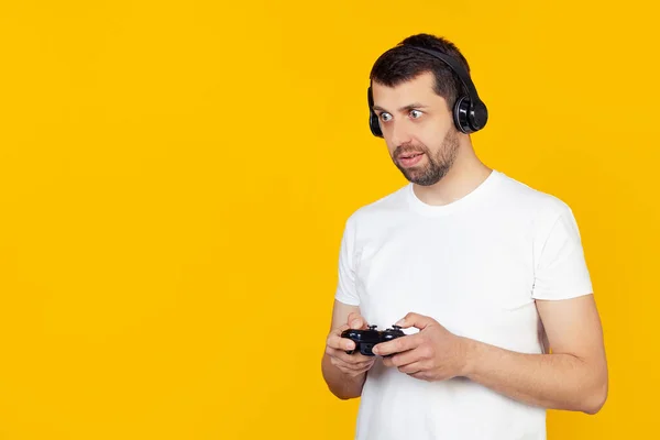 Young man with beard in white t-shirt gamer playing video games with headphones smiling and looking at the camera. Stands on isolated yellow background.