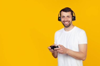 Beyaz tişörtlü sakallı genç adam video oyunları oynuyor kulaklıkla gülümsüyor ve kameraya bakıyor. İzole edilmiş sarı arkaplanda durur.