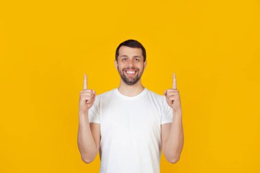 Beyaz tişörtlü sakallı genç bir adam kameraya bakıp ellerini kaldırarak mutlu bir şekilde gülümsüyor. İzole edilmiş sarı arkaplanda durur