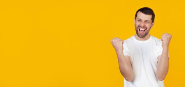 Beyaz tişörtlü sakallı genç bir adam çok mutlu ve heyecanlı, kazanan bir jest yapıyor, gülümseyerek ve başarıyla bağırarak. Kutlama kavramı izole edilmiş sarı arkaplanda duruyor