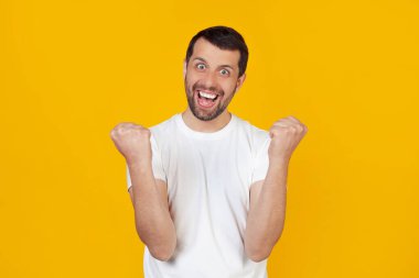 Beyaz tişörtlü sakallı genç bir adam çok mutlu ve heyecanlı, kazanan bir jest yapıyor, gülümseyerek ve başarıyla bağırarak. Kutlama kavramı izole edilmiş sarı arkaplanda duruyor