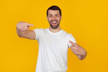 Beyaz tişörtlü sakallı genç adam mutlu ve komik bir yüzle kamerayı işaret ediyor. İyi enerji ve titreşimler. İzole edilmiş sarı arkaplanda durur.