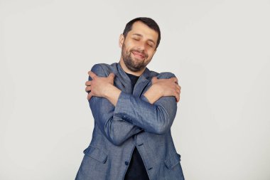 Sakallı genç işadamı mutlu ve olumlu bir şekilde kendine sarılıyor, kendine güvenerek gülümsüyor. Kendini sevme ve kendini koruma. Gri arka planda bir adamın portresi.