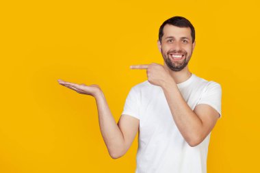 Young man with a beard in a white T-shirt is amazed and smiles at the camera, showing with his hand and pointing with his finger. Stands on isolated yellow background. clipart