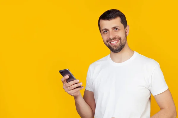 Beyaz tişörtlü, sakallı genç adam gülümseyen bir gülümseme ve gülümseyen, gülen bir akıllı telefon kullanıyor. İzole edilmiş sarı arkaplanda durur.