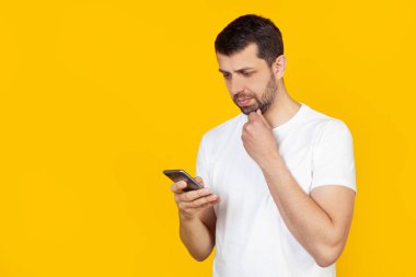 Beyaz tişörtlü sakallı genç bir adam akıllı yüzünde kendine güvenen bir ifadeyle akıllı bir telefon kullanır, ciddi düşünür. İzole edilmiş sarı arkaplanda durur.