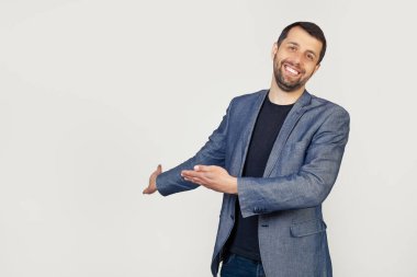 Ceketli, sakallı genç bir işadamı, içeri girmeye davet ediyor, doğal bir şekilde gülümsüyor. Gri arka planda bir adamın portresi.