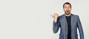 Ceketli, sakallı genç bir işadamı, şaşırmış, açık ağızlı, şaşkın bir ifadeyle işaret ediyor. Gri arka planda bir adamın portresi.