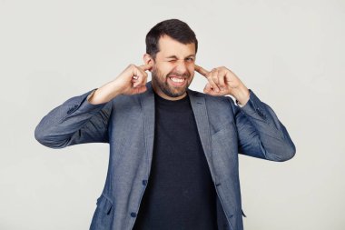 Ceketli sakallı genç bir işadamı yüksek sesli müziğin gürültüsü yüzünden yüzünde rahatsız edici bir ifadeyle parmaklarıyla kulaklarını kapatıyor. Sağır konsepti. Gri arka planda bir adam.