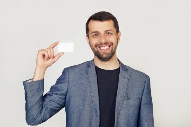 Ceketli sakallı genç bir işadamı boş bir sayfa kağıt gösteriyor. Adam gülümsüyor ve elinde bir kartvizit tutuyor. Gri arka planda bir adamın portresi.