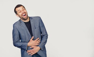 Ceketli, sakallı genç bir işadamı. Ellerini vücuduna koyduğu komik bir şakadan dolayı gülümsüyor ve kahkaha atıyor. Gri arka planda bir adamın portresi.