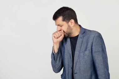 Ceketli sakallı genç bir işadamı, kendini kötü hissediyor ve soğuk algınlığı veya bronşitin bir belirtisi olarak öksürüyor. Sağlık hizmeti konsepti. Gri arka planda bir adamın portresi.