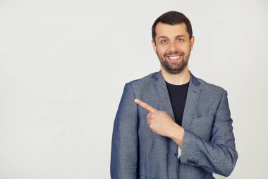 Ceketli, sakallı, neşeli, yüzünde bir gülümseme olan genç bir işadamı, mutlu ve doğal bir ifadeyle elini ve parmağını yana gösteriyor. Gri arka planda bir adamın portresi.