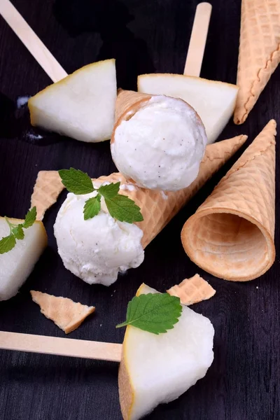 White ice cream in waffle cones. Pieces of melon around on a dark table