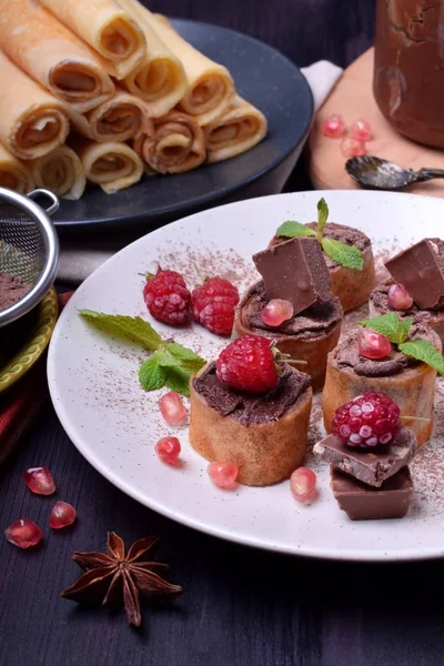 Rolled crepes with chocolate paste topped with raspberries, pomegranate, mint and chocolate pieces on a white plate