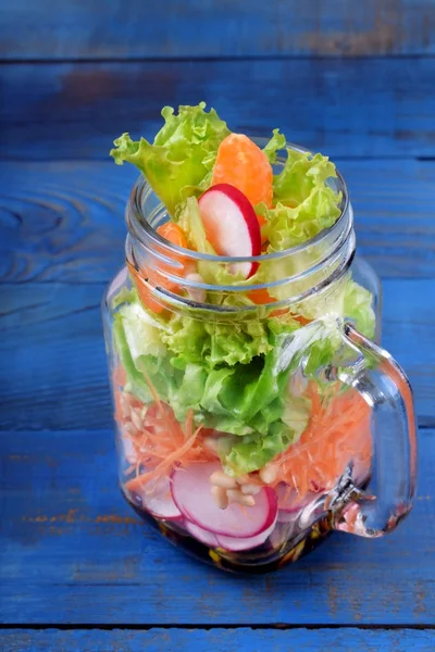 Layered salad in a jar with mandarins, radish, lettuce leaves, carrot and dressing against the blue background