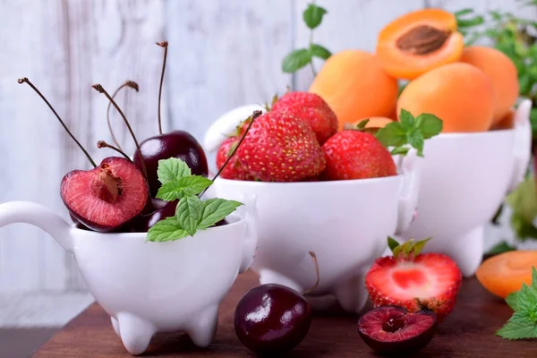 Cherries, strawberries and apricots in little white cups on the wooden table