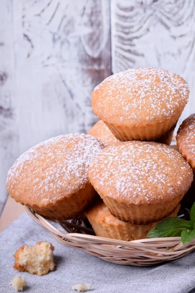 Fresh muffins topped with sugar powder in a wicker basket against the white wooden wall