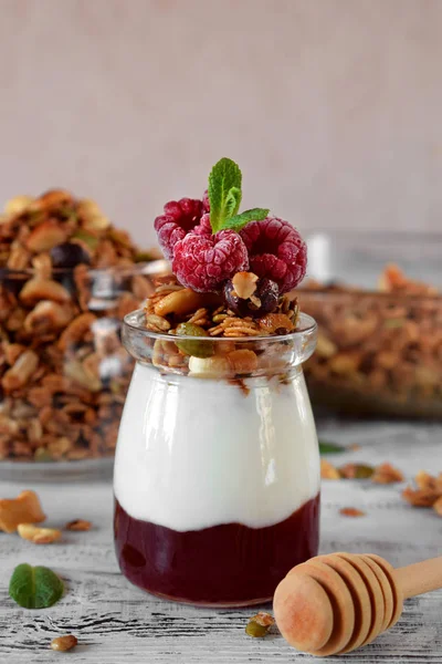 Layered breakfast with granola, yogurt and jam topped with raspberries in a glass jar on the white table