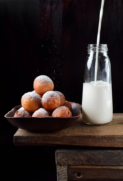 Round donuts topped with sugar powder and milk poured into glass bottle against black background