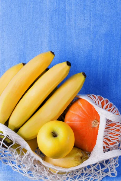 Bunch of bananas, pear, apple and small pumpkin in the mesh bag against the blue background. Copy space