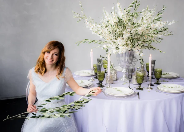 Sitting bride with olive branch in hands  and wedding decorations for banquet with sakura, candles and glass drops