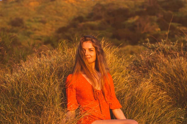 Jovem sorrindo menina loira em vestido vermelho sentado na grama — Fotografia de Stock