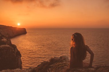 Genç bir kadın, gün batımında sahilde oturuyor.