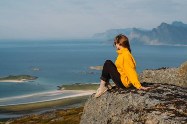 Sarı ceketli kız kayaların kenarına dikilmiş. 