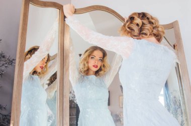 Klasik makyajlı ve saç stili güzel sarışın kız. Üçlü aynanın yanında durup kameraya bakıyor.