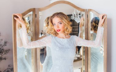 Klasik makyajlı ve saç stili güzel sarışın kız. Üçlü aynanın yanında durup kameraya bakıyor.