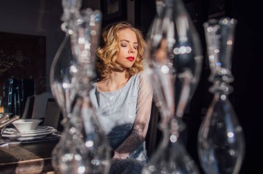 Kaliteli makyajlı, saç stili, yüzünde güneş ışınları, ön planda cam şamdanlar olan güzel bir sarışın kız.