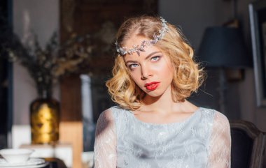 Kaliteli makyajlı, yüzünde güneş ışınları, başında mücevherler, ön planda cam şamdanlar olan güzel bir sarışın kız.