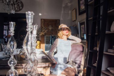 Kaliteli makyajlı, saç stili, yüzünde güneş ışınları, başında mücevherler, ön planda cam şamdanlar olan güzel bir sarışın kız.