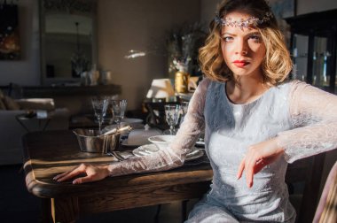 Kaliteli makyajlı, saç stili, yüzünde güneş ışınları, başında mücevherler, ön planda cam şamdanlar olan güzel bir sarışın kız.