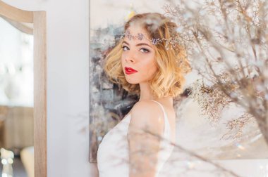 Kaliteli makyaj ve saç stili olan güzel sarışın kız aynanın yanında, başında mücevherlerle kameraya bakıyor.