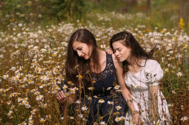 Güneşli bir günde koyu mavi ve beyaz elbiseli iki kız papatya tarlasında doğa üzerine oturuyor.