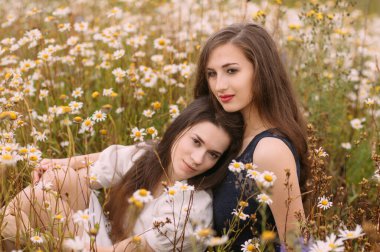 Güneşli bir günde koyu mavi ve beyaz elbiseli iki kız papatya tarlasında doğa üzerine oturuyor.