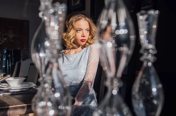 Kaliteli makyajlı, saç stili, yüzünde güneş ışınları, ön planda cam şamdanlar olan güzel bir sarışın kız.