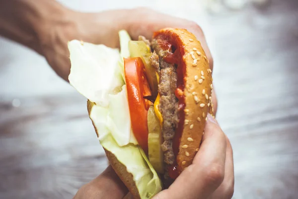 Manos sosteniendo deliciosa hamburguesa fresca en la mesa de madera vista superior . — Foto de Stock