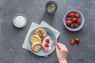 Süzme peynir kahvaltı veya gri arka plan üzerinde öğle yemeği için ekşi krema ve çilek ile krep. Sağlıklı gıda kavramı. Kadın eli bir çatal tutar. Düz yatıyordu