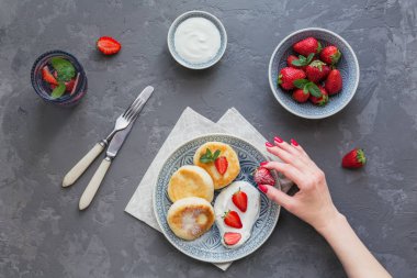 Süzme peynir kahvaltı veya gri arka plan üzerinde öğle yemeği için ekşi krema ve çilek ile krep. Sağlıklı gıda kavramı. Kadın eli bir dut tutar. Üstten görünüm