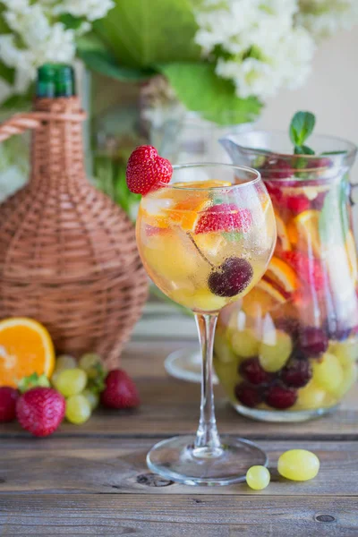 Geleneksel yaz içki beyaz köpüklü şarap sangria. Homemade Meyve sangria şampanya, çilek, portakal ve üzüm ile yenileniyor. Bir sürahi ve malzemeler ile ahşap rustik masaya
