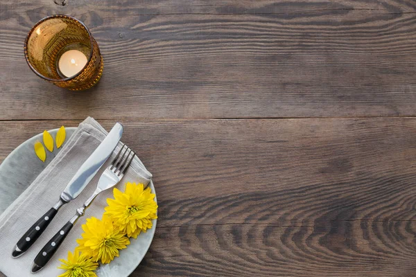 Rustic table setting with linen napkin, cutlery, ceramic plates, yellow glasses and yellow flowers on dark wooden table. Holiday table decoration. Romantic dinner.