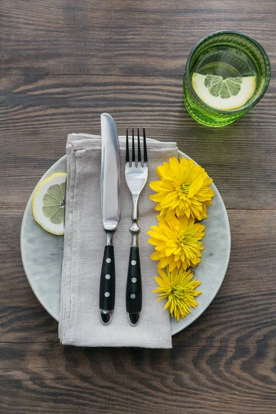 Rustic table setting with linen napkin, cutlery, ceramic plates, yellow glasses and yellow flowers on dark wooden table. Holiday table decoration. Romantic dinner.