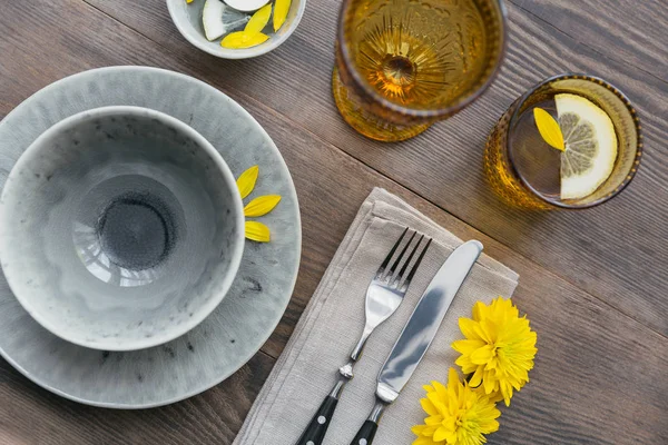 Rustic table setting with linen napkin, cutlery, ceramic plates, yellow glasses and yellow flowers on dark wooden table. Holiday table decoration. Romantic dinner.