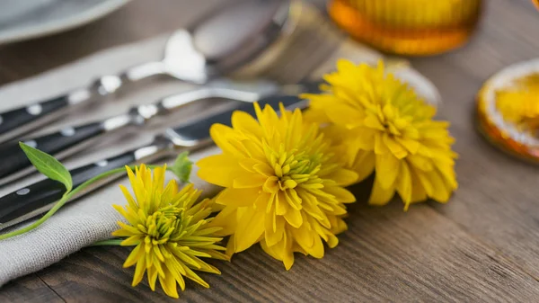 Rustic table setting with linen napkin, cutlery, ceramic plates, yellow glasses and yellow flowers on dark wooden table. Holiday table decoration. Romantic dinner.